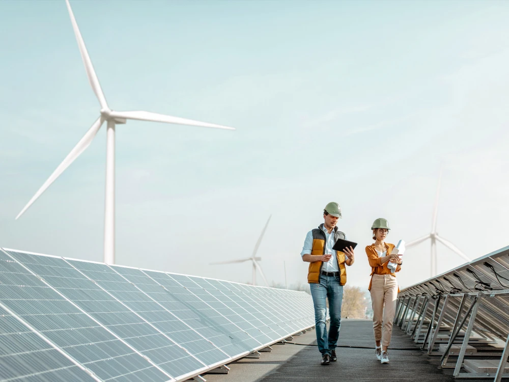 Two engineers in safety helmets reviewing documents at a solar farm with wind turbines in the background