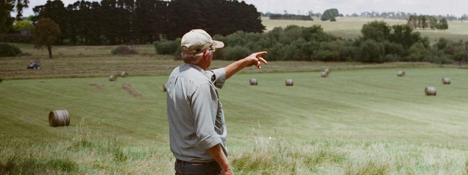 Ein älterer Mann mit Kappe zeigt auf eine weite Landschaft mit Strohballen