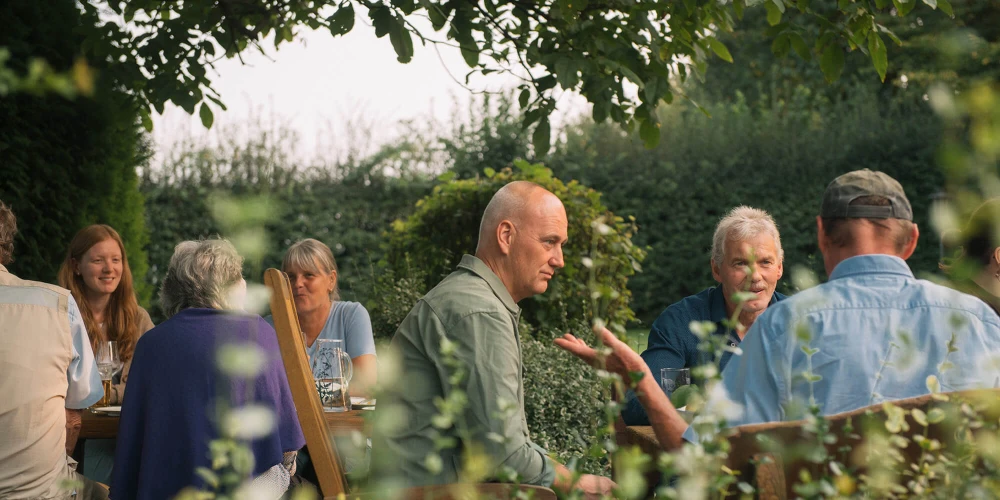 Gruppe von Menschen unterschiedlichen Alters sitzt im Freien um einen Tisch herum und unterhält sich in einem Garten