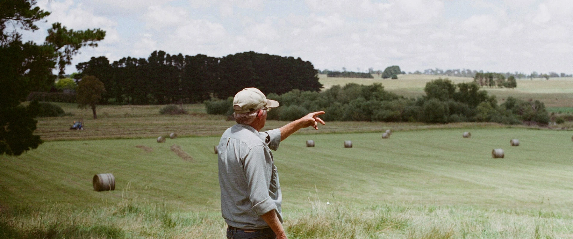 Elderly man with a cap pointing at a field with hay bales