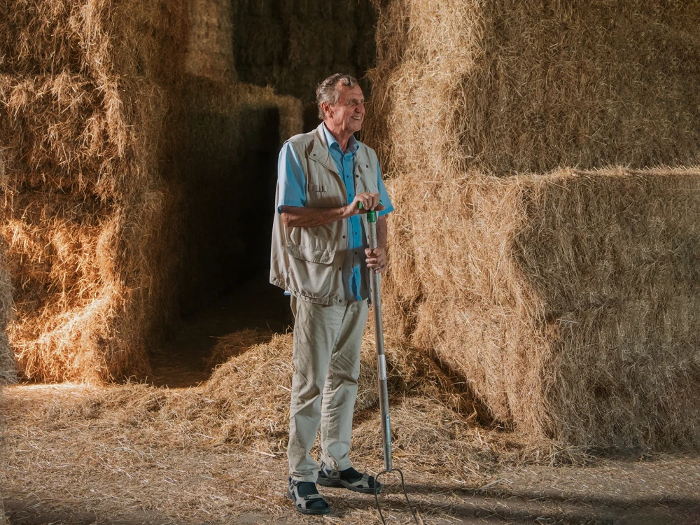 Älterer Mann steht in einer Scheune zwischen großen Heuballen und hält eine Mistgabel