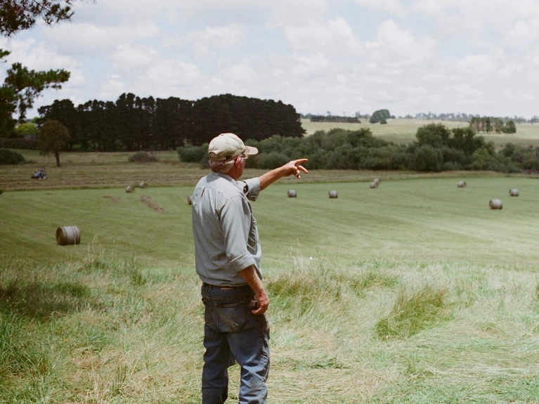 Older man wearing a cap pointing at a rural landscape with hay bales