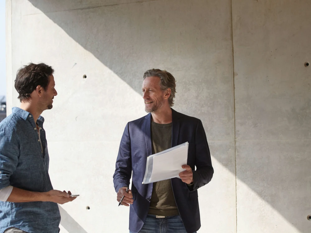 Two men talking in front of a concrete wall, one holding documents