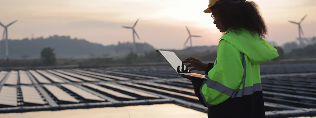Engineer in safety gear checking data on a laptop in front of a solar field and wind turbines at sunset