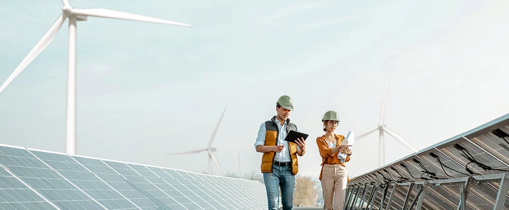 Two engineers in safety helmets walking among solar panels and wind turbines while looking at a document