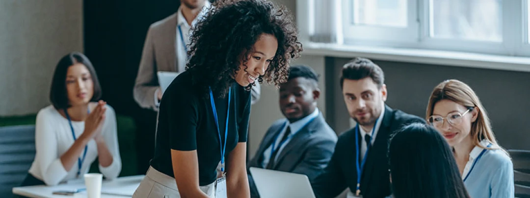 Young curly-haired woman with a lanyard standing and presenting on a laptop to seated colleagues in a conference room