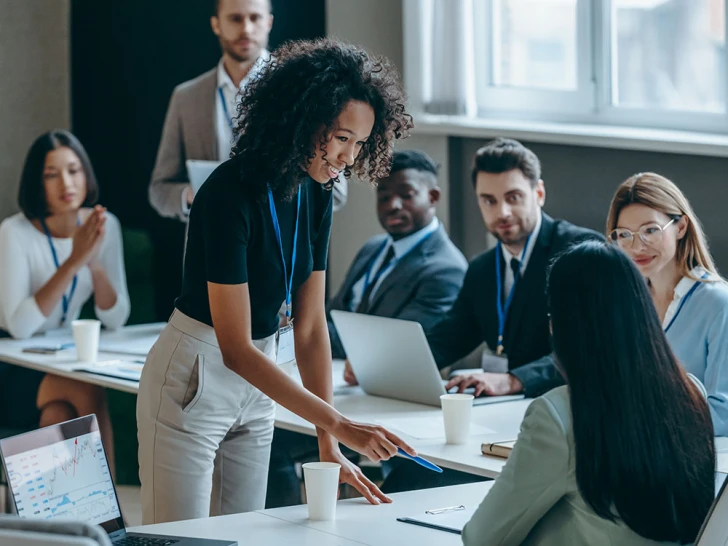 A young woman presenting in a meeting room in front of a group of businesspeople sitting at a table and listening