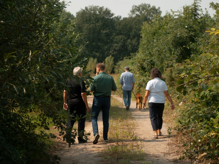 Several people walking down a narrow path surrounded by green trees, one person is leading a dog on a leash