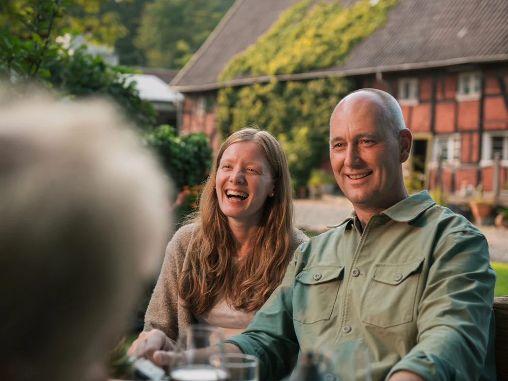 Lachendes Paar sitzt an einem Tisch im Freien vor einem traditionellen Haus