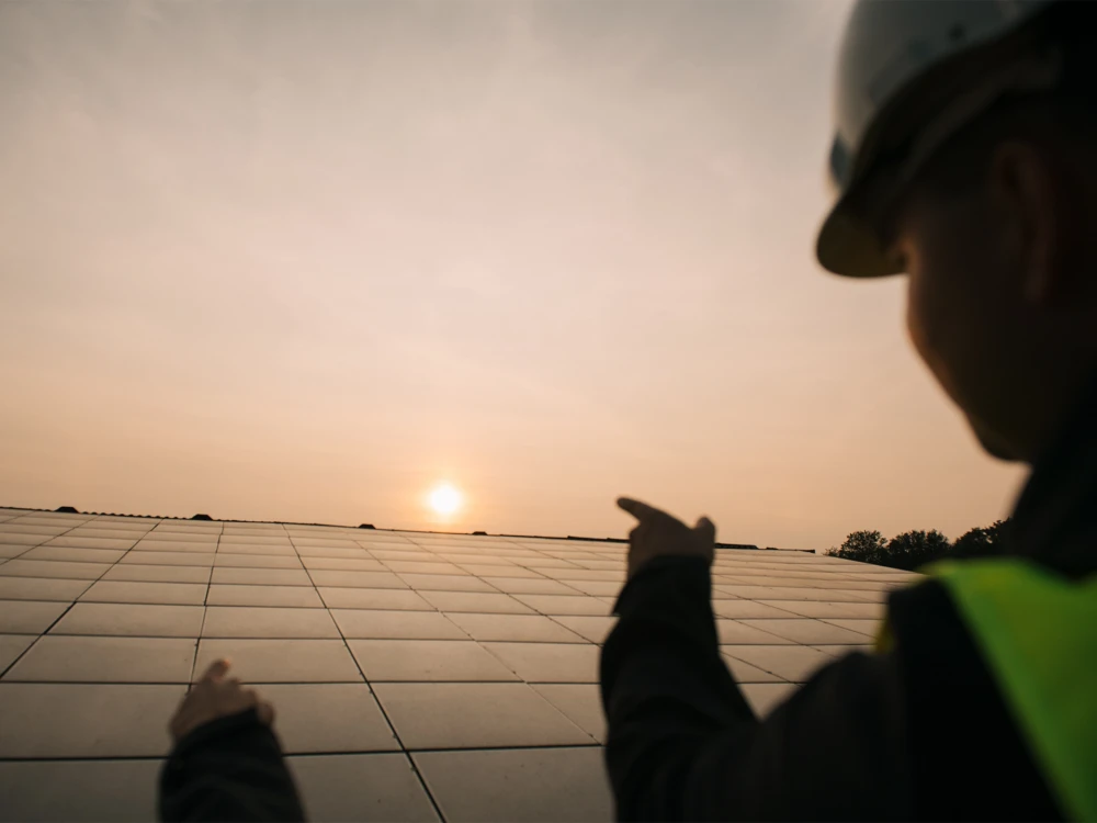 Person in work attire and helmet pointing at solar panels at sunset