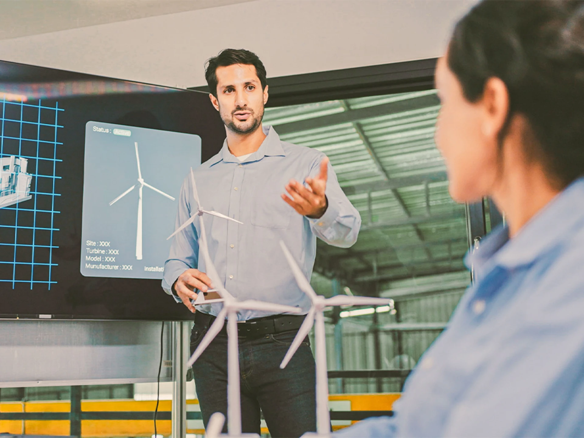 Man presenting wind turbine models in front of a digital display during a discussion with a female colleague