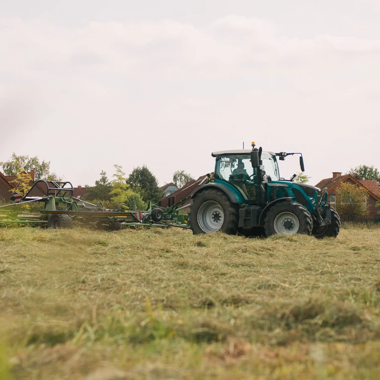 Grüner Traktor auf einem Feld beim Mähen von Gras, im Hintergrund Häuser
