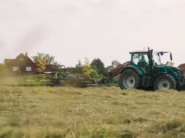 Grüner Traktor auf einem Feld beim Mähen, unscharfer Vordergrund, ländliche Szenerie im Hintergrund