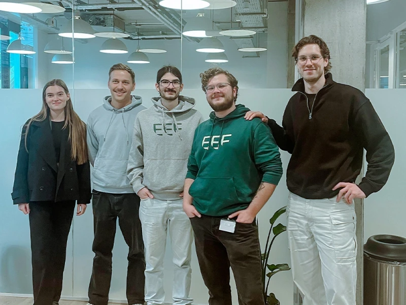 Fünf junge Erwachsene stehen lächelnd in einem modernen Büro