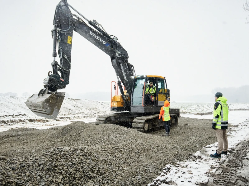 An excavator operating at a construction site in the snow, with two people in safety clothing watching