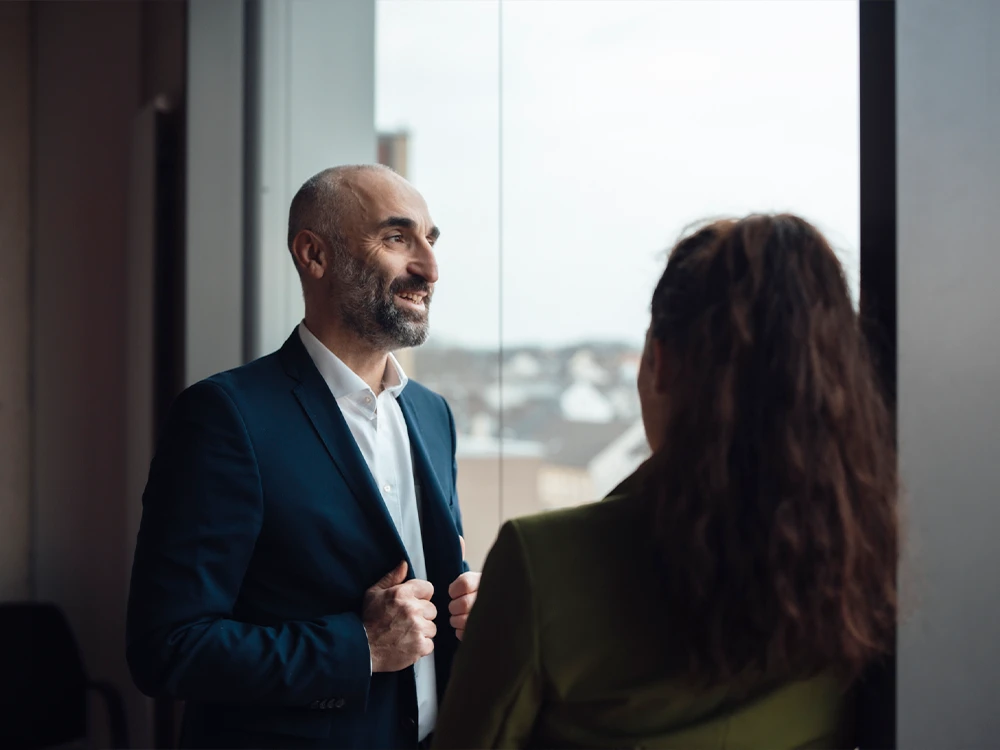 Bald man in a suit smiling and talking to a woman with her back turned to him