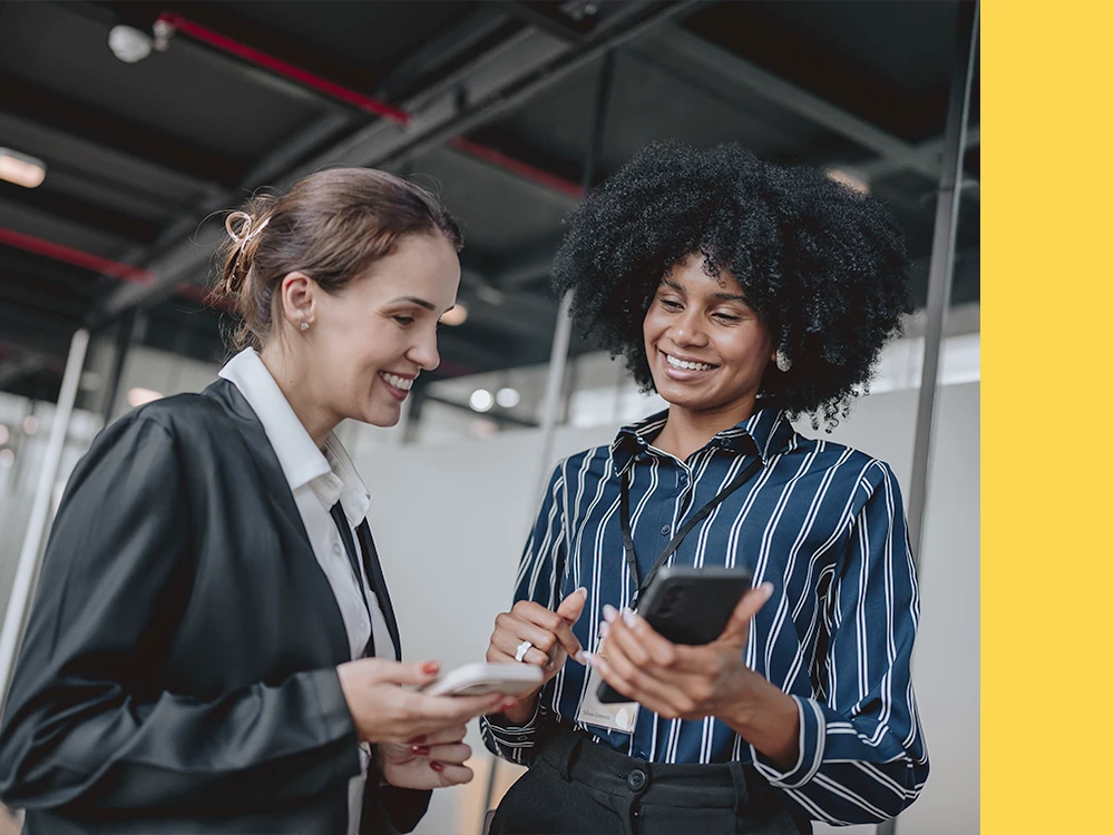 Two businesswomen, one with Afro-textured hair, looking at a smartphone together in a modern office setting