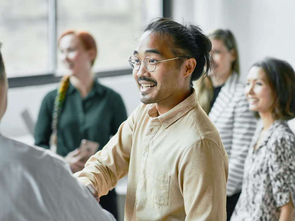 Ein Mann mit Brille und Hemd gibt in einem Büro mit Kollegen im Hintergrund einem anderen Mann die Hand