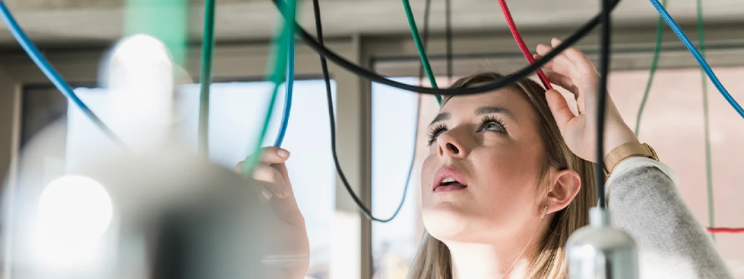 Young woman looking thoughtfully at several colorful cables hanging above her in an office