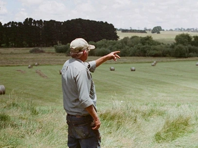 Älterer Mann auf einem Feld, der in die Ferne zeigt, umgeben von Heuballen