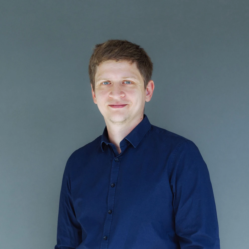 Porträt eines lächelnden jungen Mannes mit kurzen blonden Haaren, der ein dunkelblaues Hemd trägt und vor einem grauen Hintergrund steht