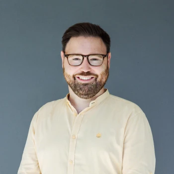 Portrait of a smiling man with a beard and glasses wearing a light yellow shirt against a gray background