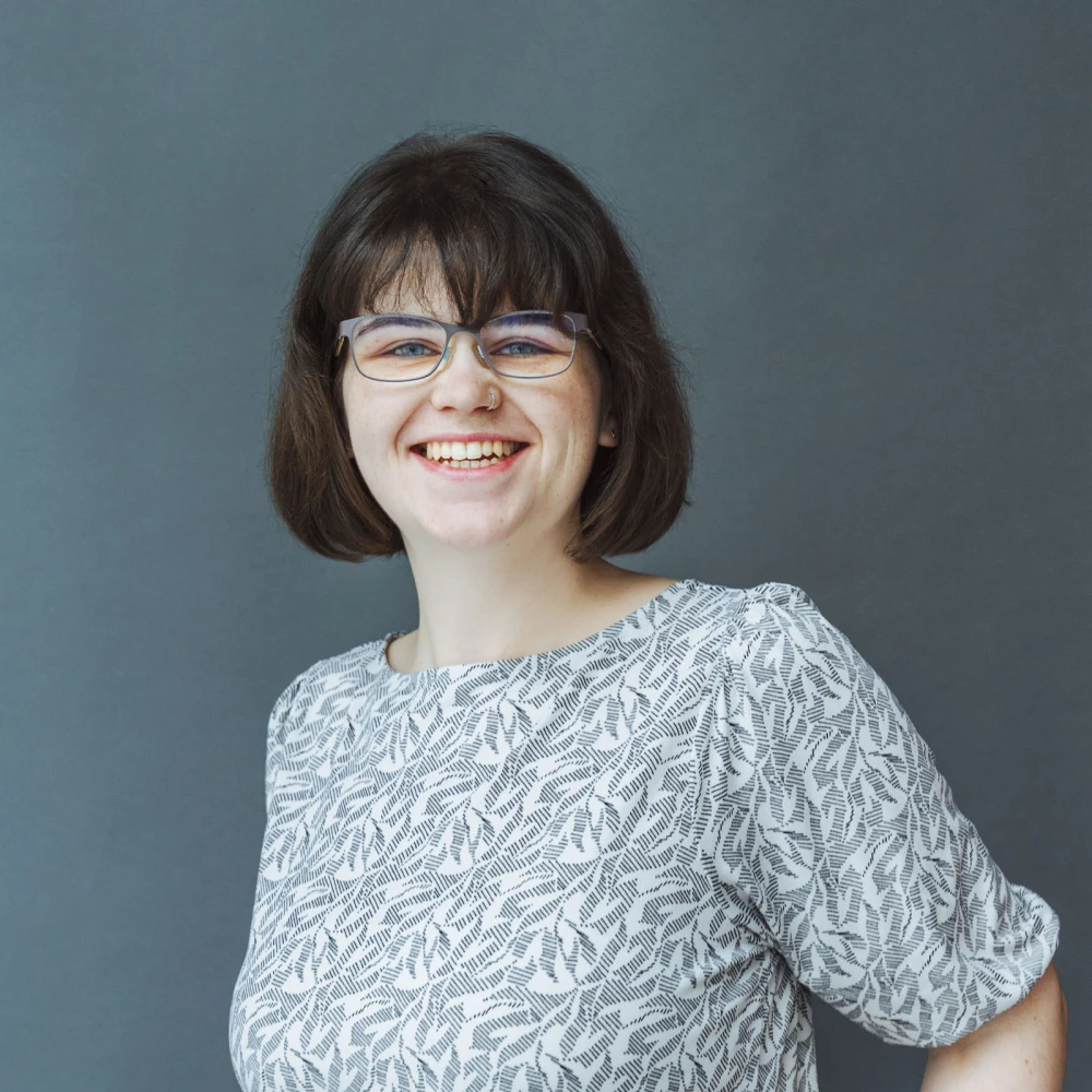 Porträt einer lächelnden Frau mit Brille und kurzem Haar vor einem dunkelgrauen Hintergrund