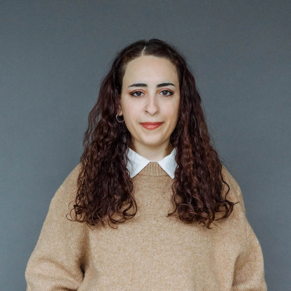 Porträt einer Frau mit lockigem braunem Haar und einem beigen Pullover vor grauem Hintergrund
