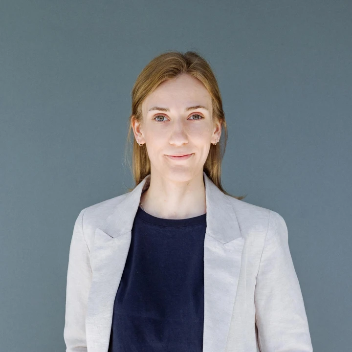 Porträt einer Frau mit blonden Haaren in einem grauen Blazer und dunkelblauem Oberteil, vor einem grauen Hintergrund