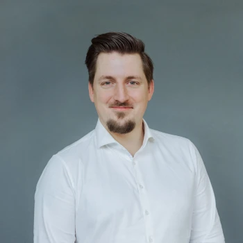Portrait of a smiling man with dark hair and beard wearing a white shirt against a grey background