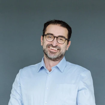 Smiling man with glasses and gray beard wearing a light blue shirt against a gray background