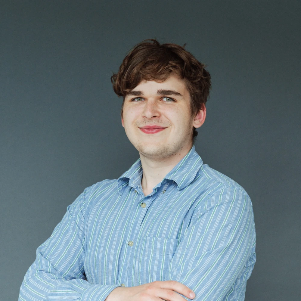 Porträt eines lächelnden jungen Mannes mit dunkelblondem Haar und blau gestreiftem Hemd vor grauem Hintergrund