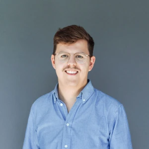 Porträt eines lächelnden Mannes mit Brille und blauem Hemd vor grauem Hintergrund