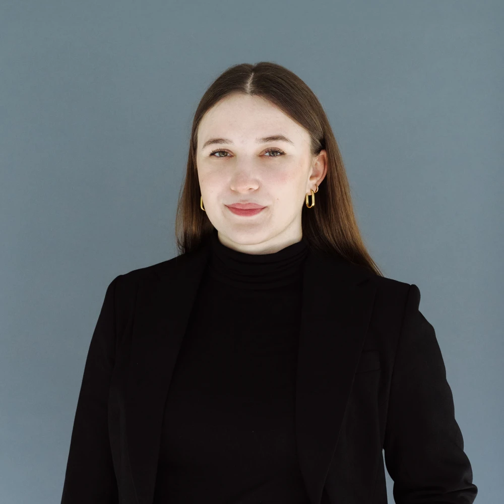 Porträt einer jungen Frau mit langen Haaren, die eine schwarze Jacke und einen Rollkragen trägt, vor einem grauen Hintergrund