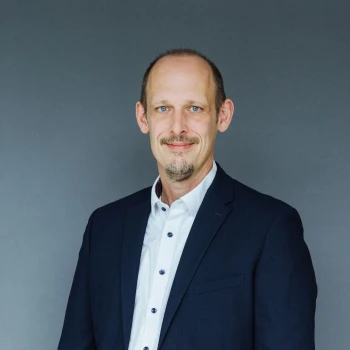Portrait of a smiling bald man with a beard in a dark blue suit against a gray background