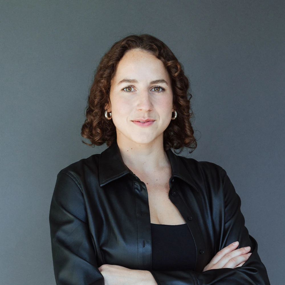 Porträt einer lächelnden Frau mit lockigen braunen Haaren und schwarzer Kleidung, die vor einem grauen Hintergrund steht