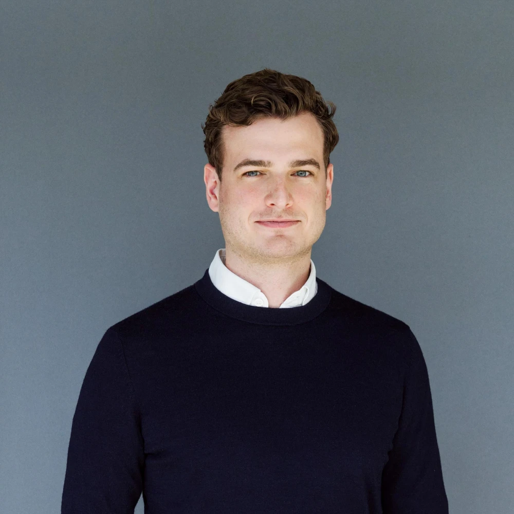 Porträt eines lächelnden Mannes mit dunklem Pullover und weißem Hemd vor grauem Hintergrund