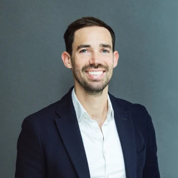Portrait of a smiling man with dark hair and a beard, wearing a dark blue jacket and a white shirt