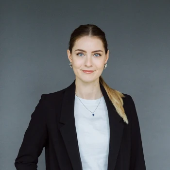 Portrait of a young woman with tied-back blonde hair wearing a black jacket and white shirt, against a grey background