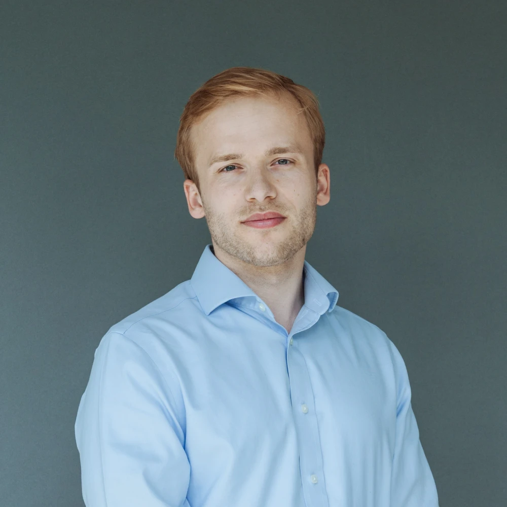 Porträt eines lächelnden Mannes mit hellblondem Haar und blauem Hemd vor grauem Hintergrund