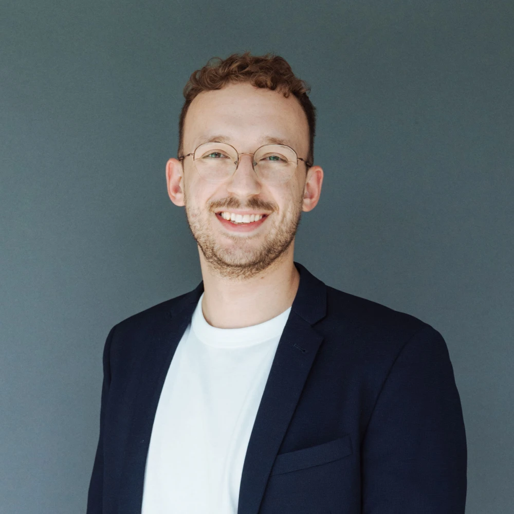 Lächelnder junger Mann mit Brille und Bart, trägt ein weißes T-Shirt und einen dunkelblauen Blazer vor einem grauen Hintergrund