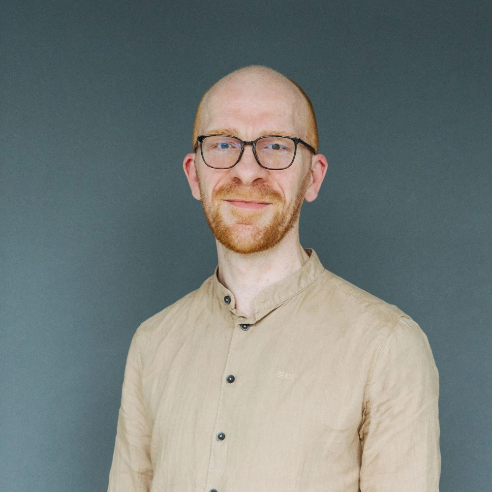 Porträt eines lächelnden kahlen Mannes mit Brille und beigem Hemd vor grauem Hintergrund