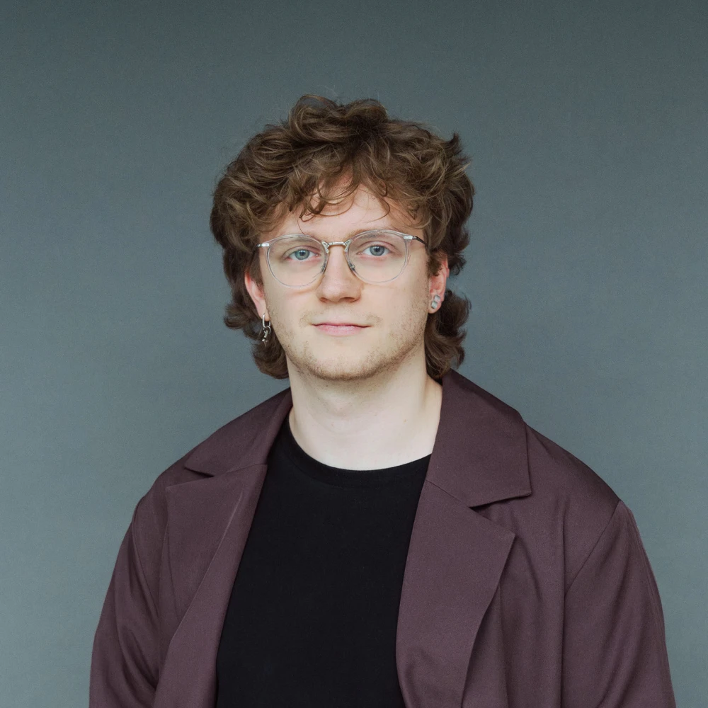 Porträt eines jungen Mannes mit lockigem Haar und Brille, gekleidet in einem dunklen T-Shirt und einem braunen Mantel vor grauem Hintergrund