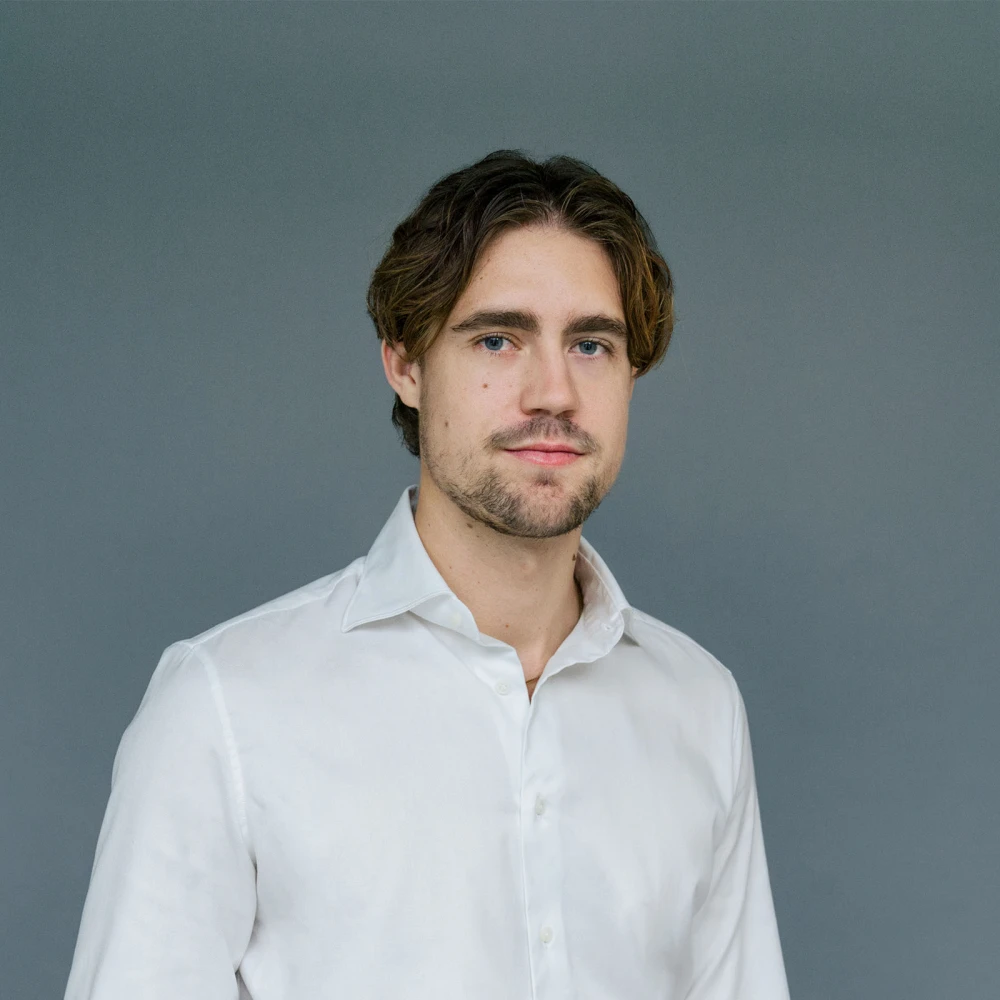 Porträt eines jungen Mannes mit mittellangem, gewelltem Haar und weißem Hemd vor grauem Hintergrund