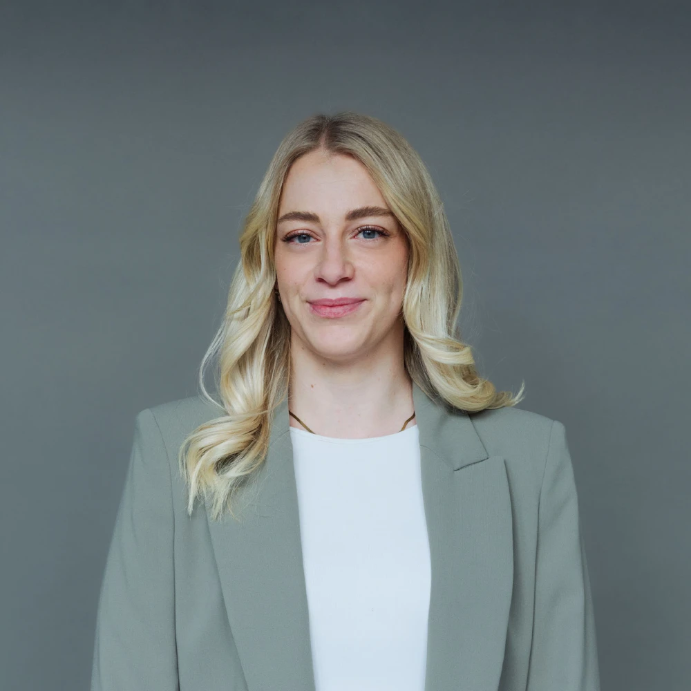 Porträt einer lächelnden blonden Frau in einem hellgrauen Blazer vor grauem Hintergrund