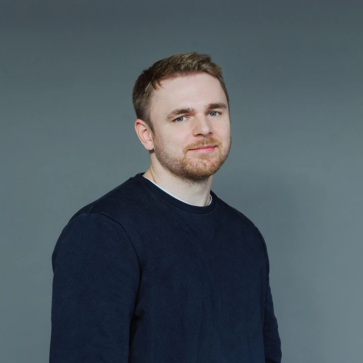 Porträt eines lächelnden Mannes mit kurzen blonden Haaren und dunkelblauem Pullover vor grauem Hintergrund