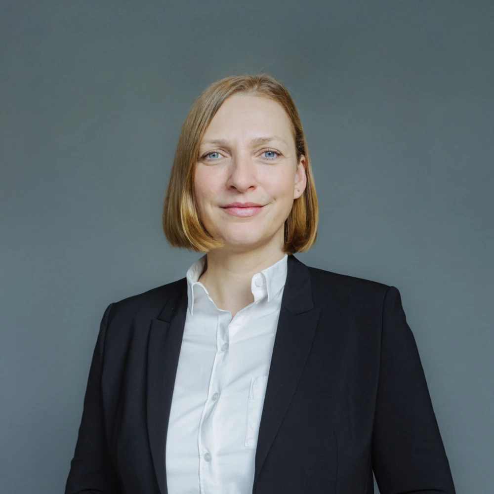 Porträt einer lächelnden Frau mit mittellangem blondem Haar in einem schwarzen Anzug und weißem Hemd vor grauem Hintergrund